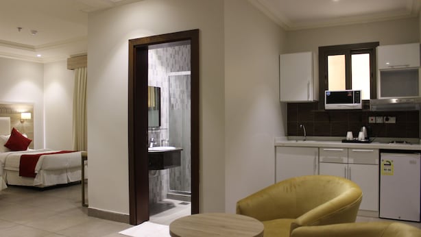 Een hotelkamer met een bed, een gele fauteuil, een klein rond tafeltje, een kitchenette met een magnetron en een koelkast, en een zichtbare badkamer door een geopende deur.
