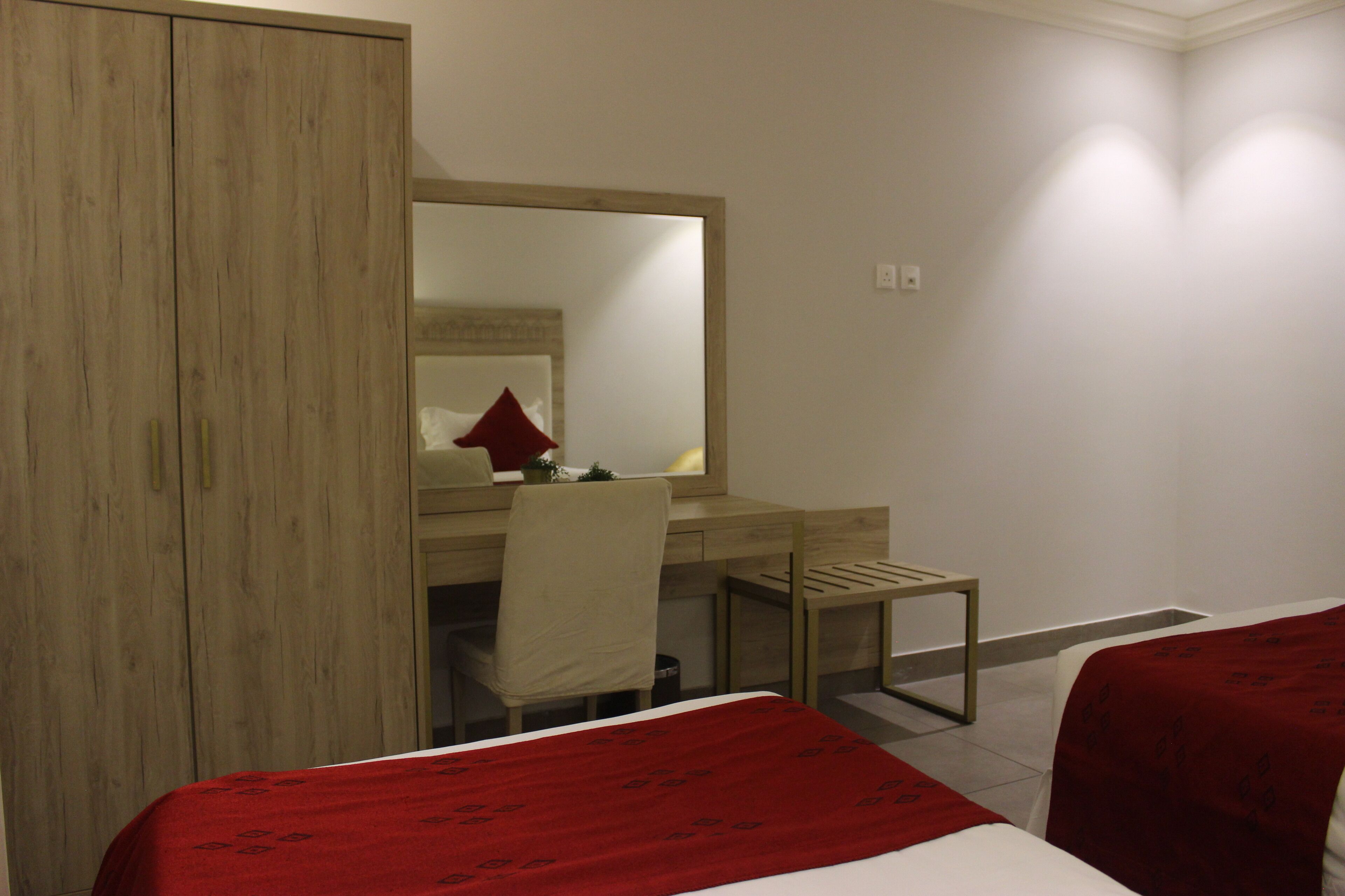Une chambre d’hôtel avec une armoire en bois, un bureau avec une chaise, un lit recouvert d’une couverture rouge et un grand miroir.