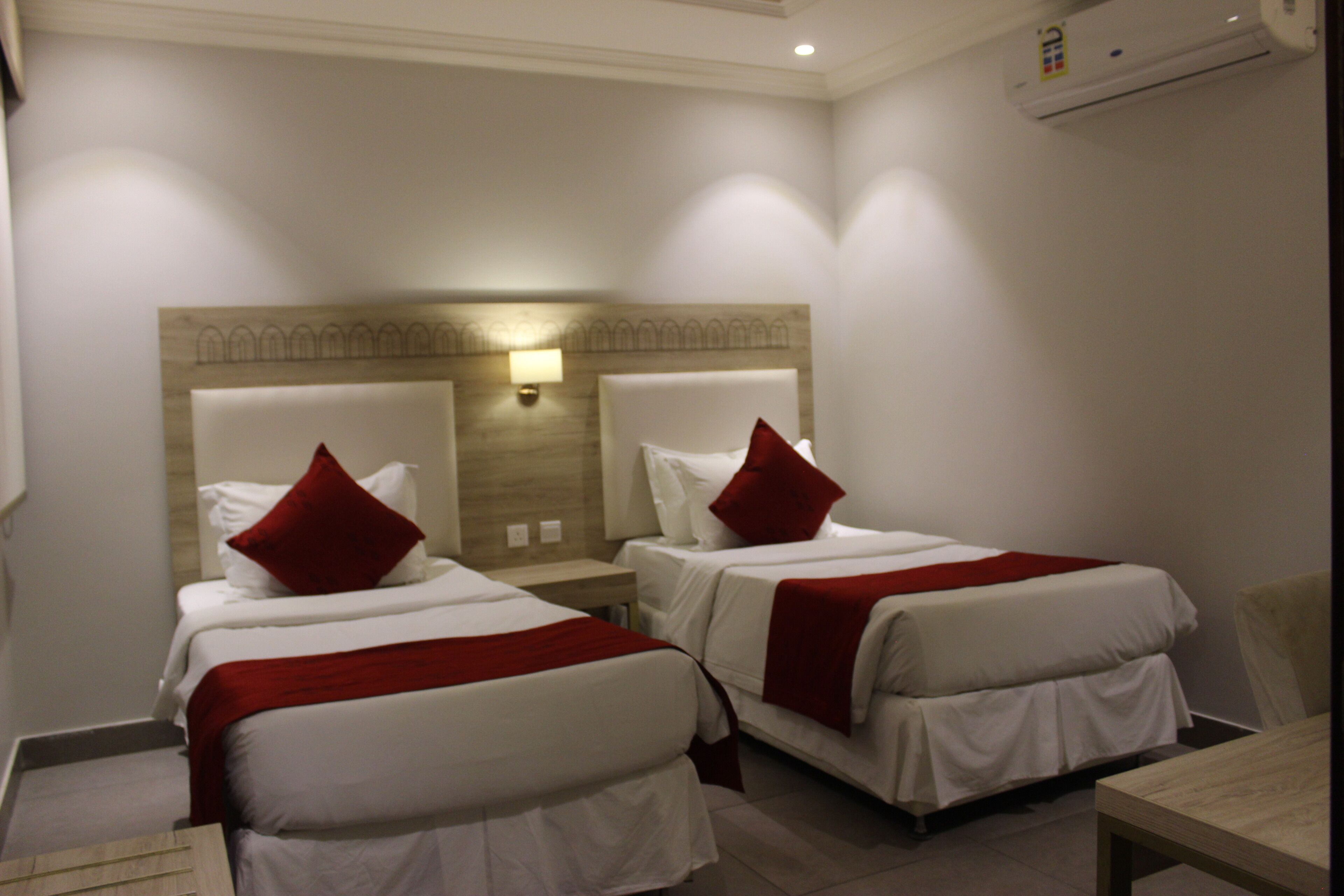 A hotel room with two single beds, each with a red pillow and white bedding. There is a wooden headboard with a light fixture above the beds.
