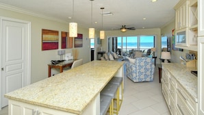A kitchen with a long island counter, a dining area with a blue patterned sofa, and a view of the ocean through large windows. - Siesta Key Stunner Endless Gulf Views from Your 2 Bed 2 Bath Top Floor Escape (Siesta Key)