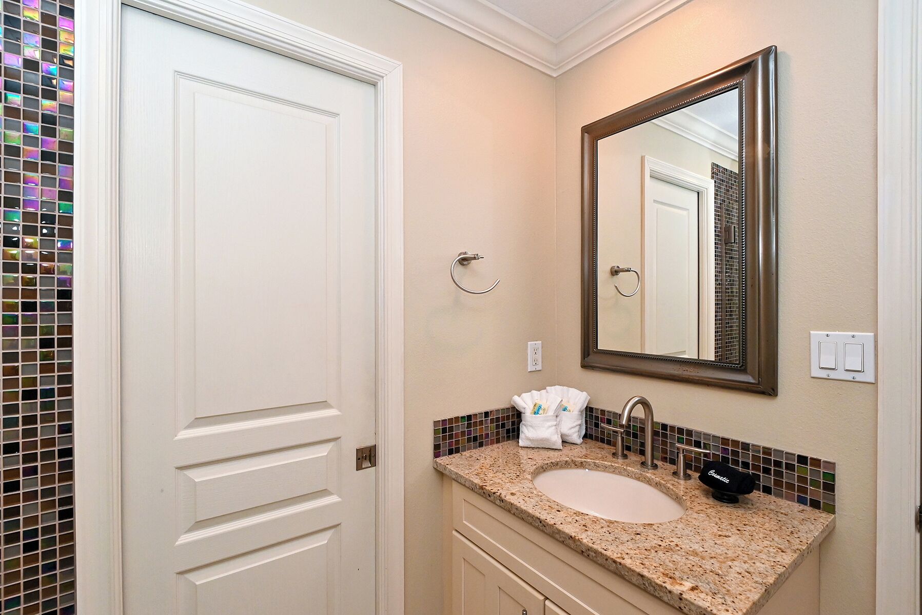 Un baño con una puerta blanca, un lavamanos con encimera de mármol, un espejo y un perchero para toallas.