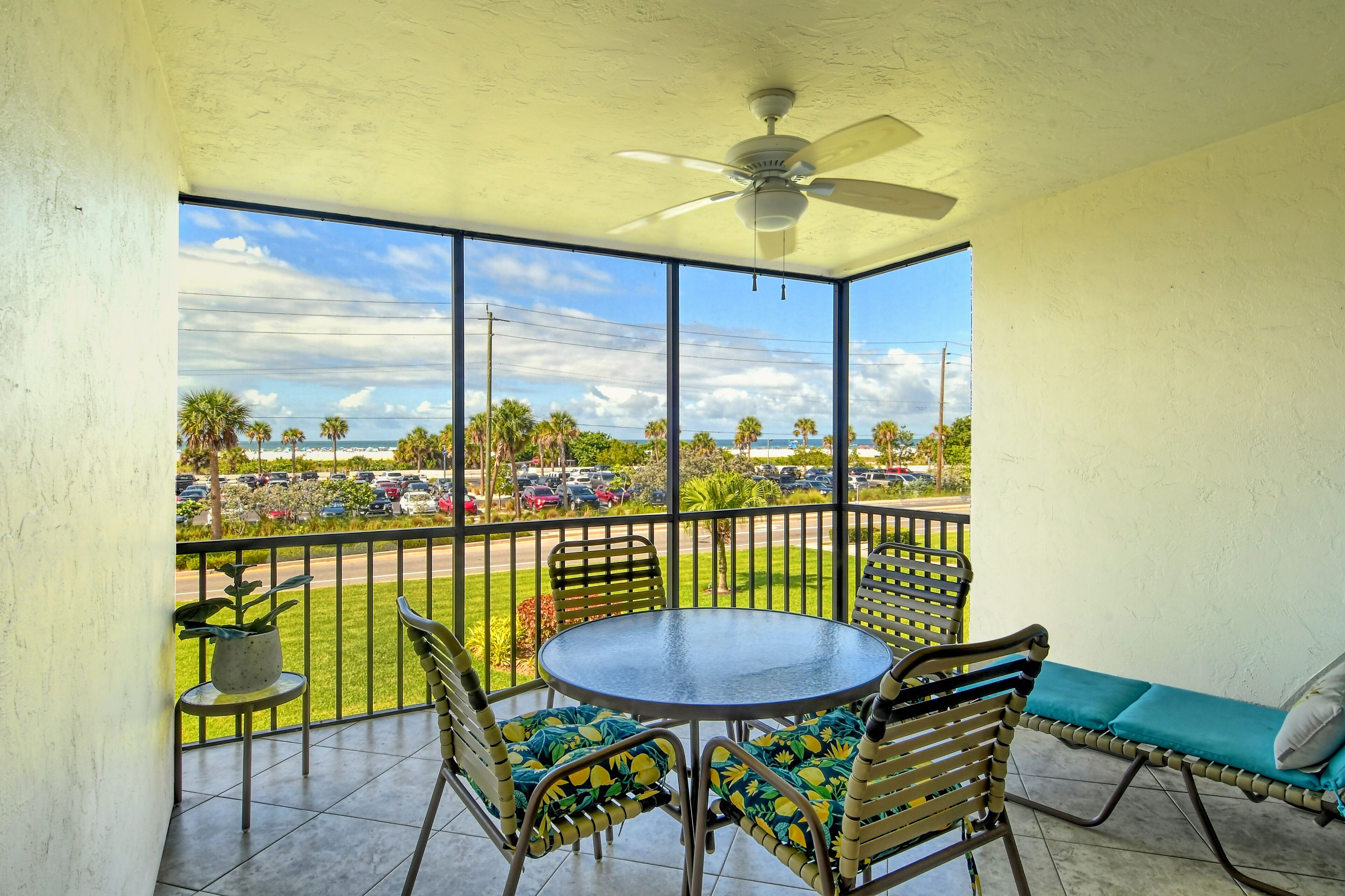 A balcony with a table and chairs, a ceiling fan, and a view of a parking lot and palm trees.
