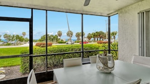 A dining area with a table and chairs, large windows with a view of the beach and palm trees, and a ceiling fan. - Serene 1st floor 2 Bed 2 Bath Siesta Key Steps to Beach (Siesta Key)