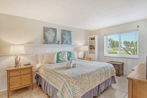 Un dormitorio con una cama grande, dos mesitas de noche, un aparador, una ventana con persianas y una pared decorada con dos cuadros.