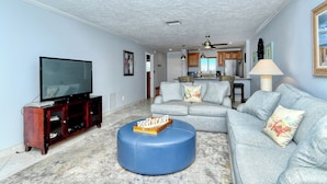 A living room with a television, sofa, ottoman, and kitchen in the background. - Siesta Key Beach 2 Bed 2 Bath with Gulf Views Steps to Beach (Siesta Key)