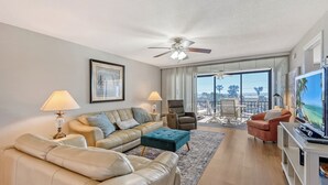 A modern living room with a sofa, ottoman, and armchairs, a ceiling fan, and a balcony with a view.