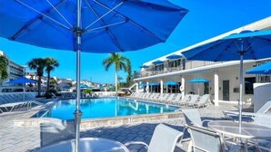 A pool area with lounge chairs and umbrellas, a clear blue sky, and a building with balconies.