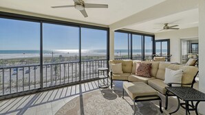 A spacious balcony with a sofa and a small table, large windows offering a view of the beach and parking area.