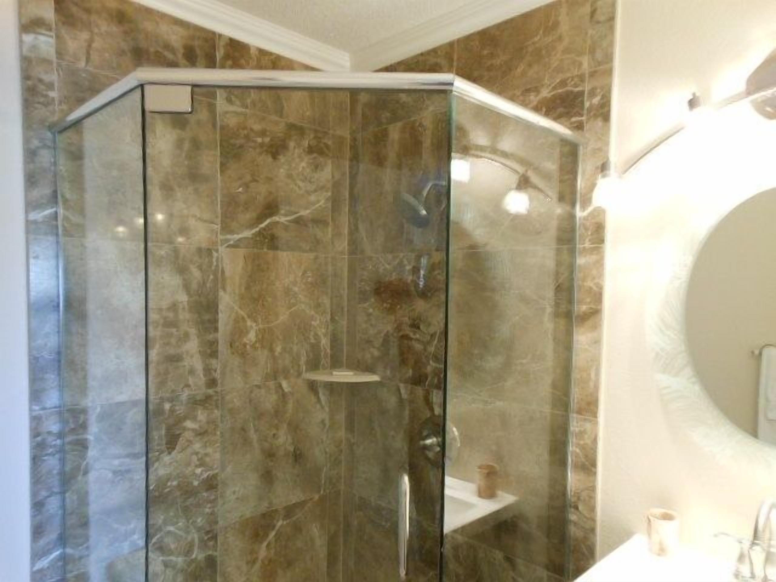 Un baño con ducha envuelta en vidrio, paredes de mármol y un espejo redondo.