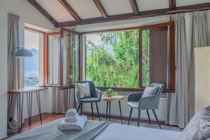 Ein Zimmer mit einem Bett, zwei Stühlen, einem kleinen Tisch mit Pflanze und Blick auf Grünflächen und Berge durch große Fenster.