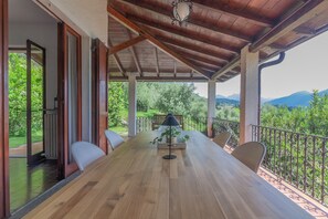 Ein überdachter Essbereich im Freien mit einem Holztisch und -stühlen, einem Blumentopf und Blick auf Grünflächen und Berge.