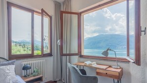 Quarto com cama, escrivaninha, cadeira, radiador e janelas grandes com cortinas.