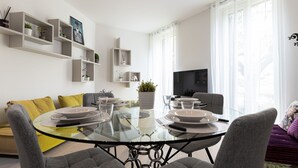 Una sala moderna con una mesa de comedor de vidrio preparada para cuatro personas, rodeada de sofás grises y un cojín amarillo. Se ven estanterías con objetos decorativos y un televisor.