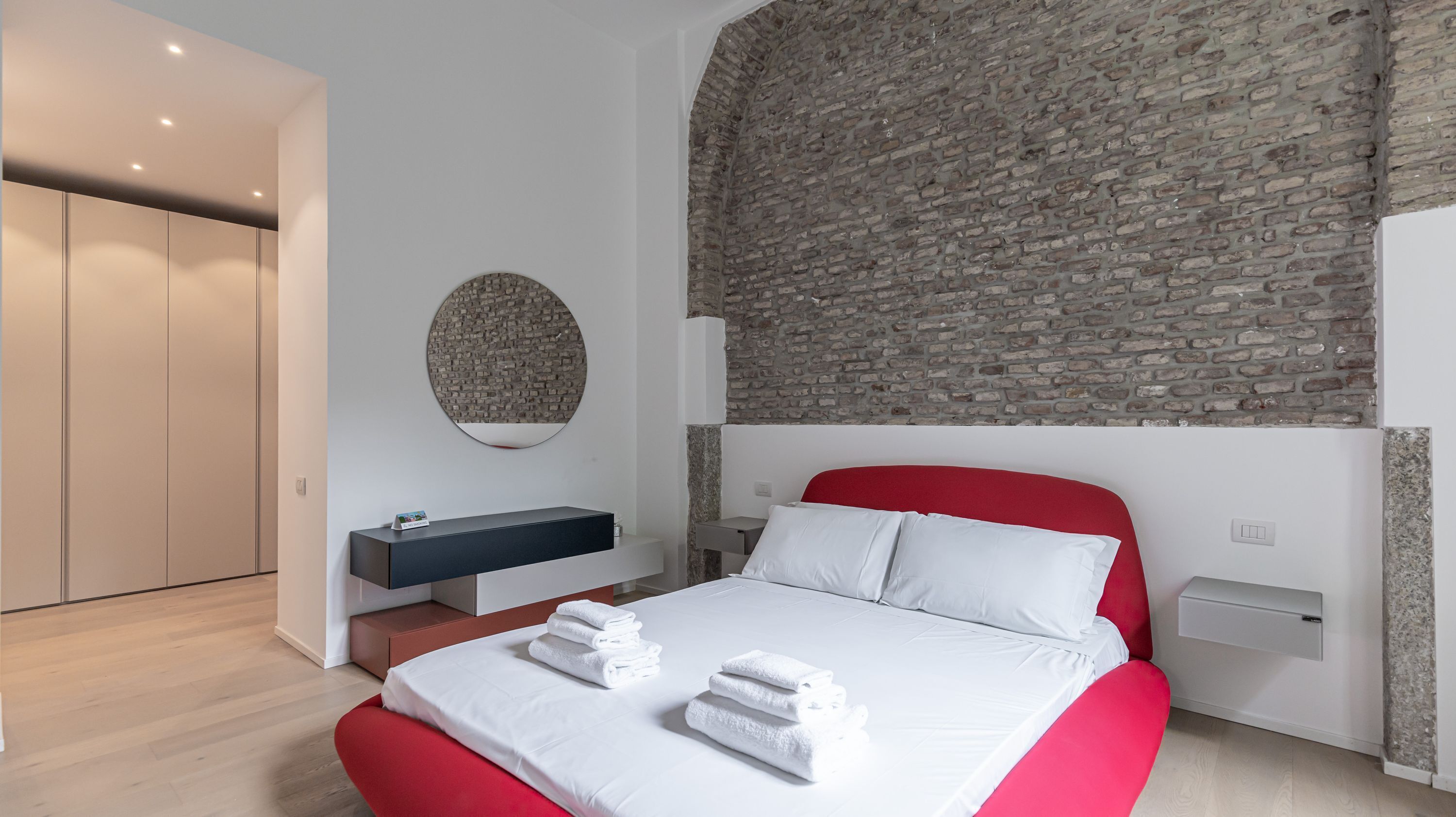 A modern bedroom with a brick wall, a round mirror, a bed with red headboard, and a bedside table.