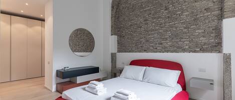 A modern bedroom with a brick wall, a round mirror, a bed with red headboard, and a bedside table.