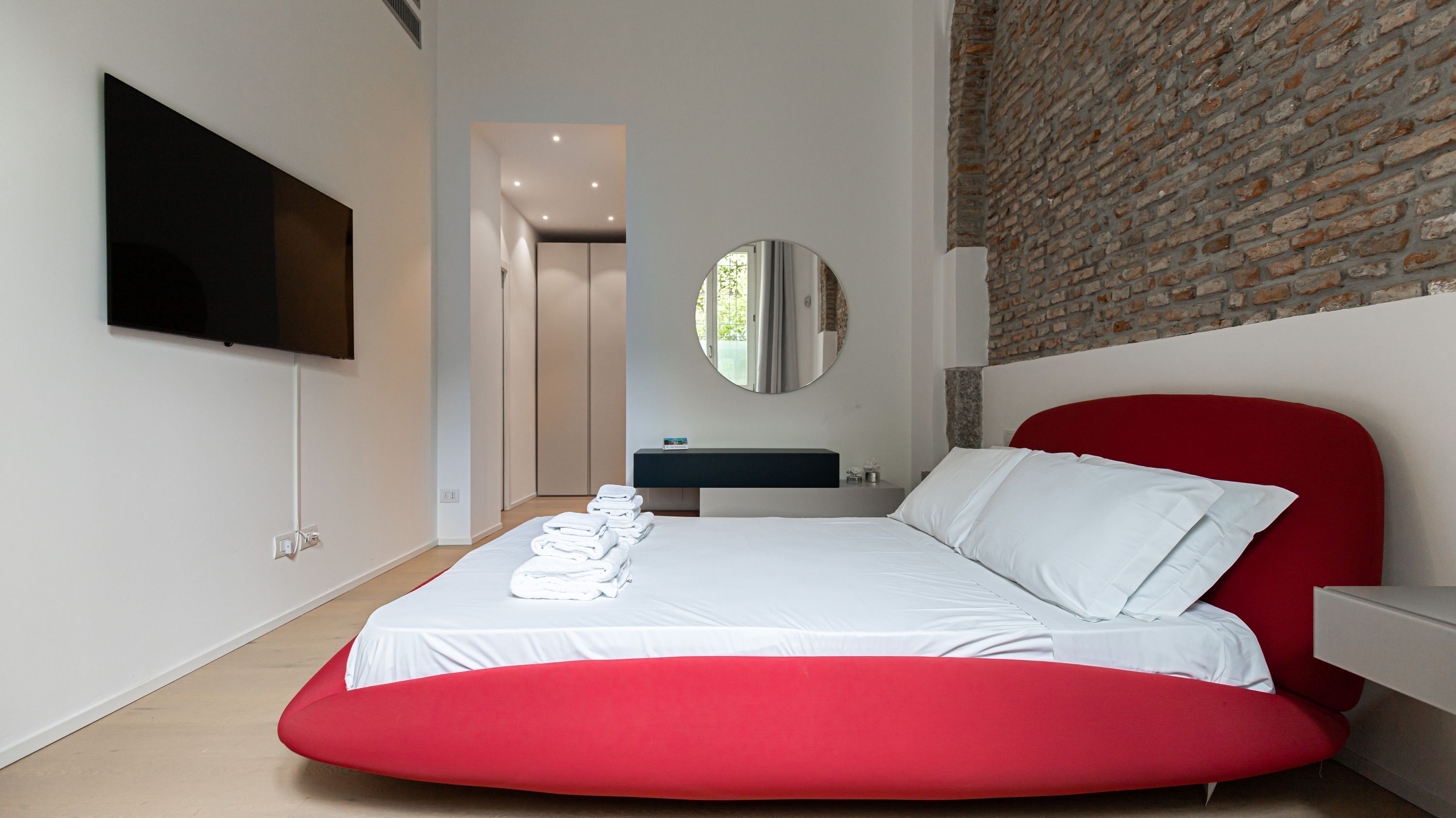 A modern bedroom with a large bed, a red accent wall, a round mirror, and a flat-screen TV mounted on the wall.