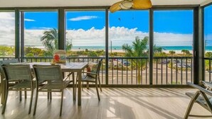 A balcony with a view of the beach and ocean, a table with chairs, and a ceiling fan.