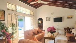 A spacious living room with a beige sofa, a coffee table, and a TV mounted on the wall. There is a large window with blinds, a ceiling fan, and a door leading outside. - Jamaica Dream Villa - Iron Shore Escape (Montego Bay)
