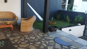Un rincón exterior acogedor con mobiliario de mimbre, suelo de baldosas de piedra y un pequeño jardín con plantas y una fuente.