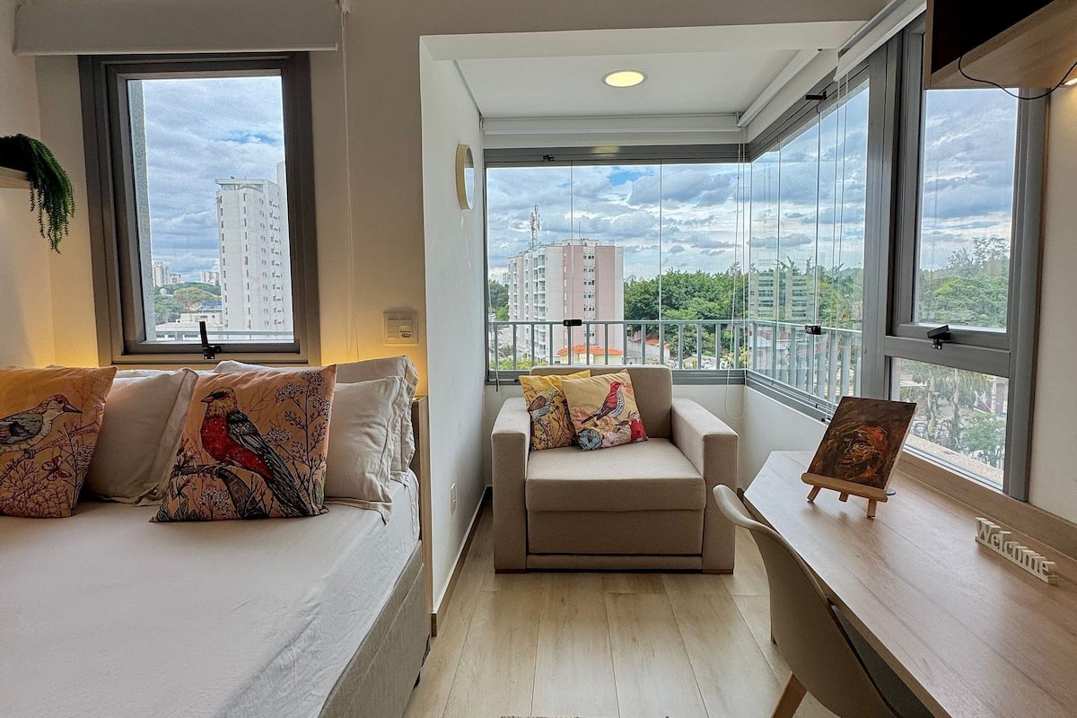 A modern apartment with a bed, a sofa, a desk, and large windows offering a city view.