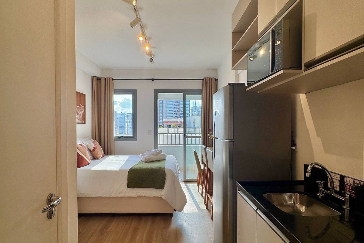 A modern hotel room with a bed, a kitchenette, and a balcony with a city view.