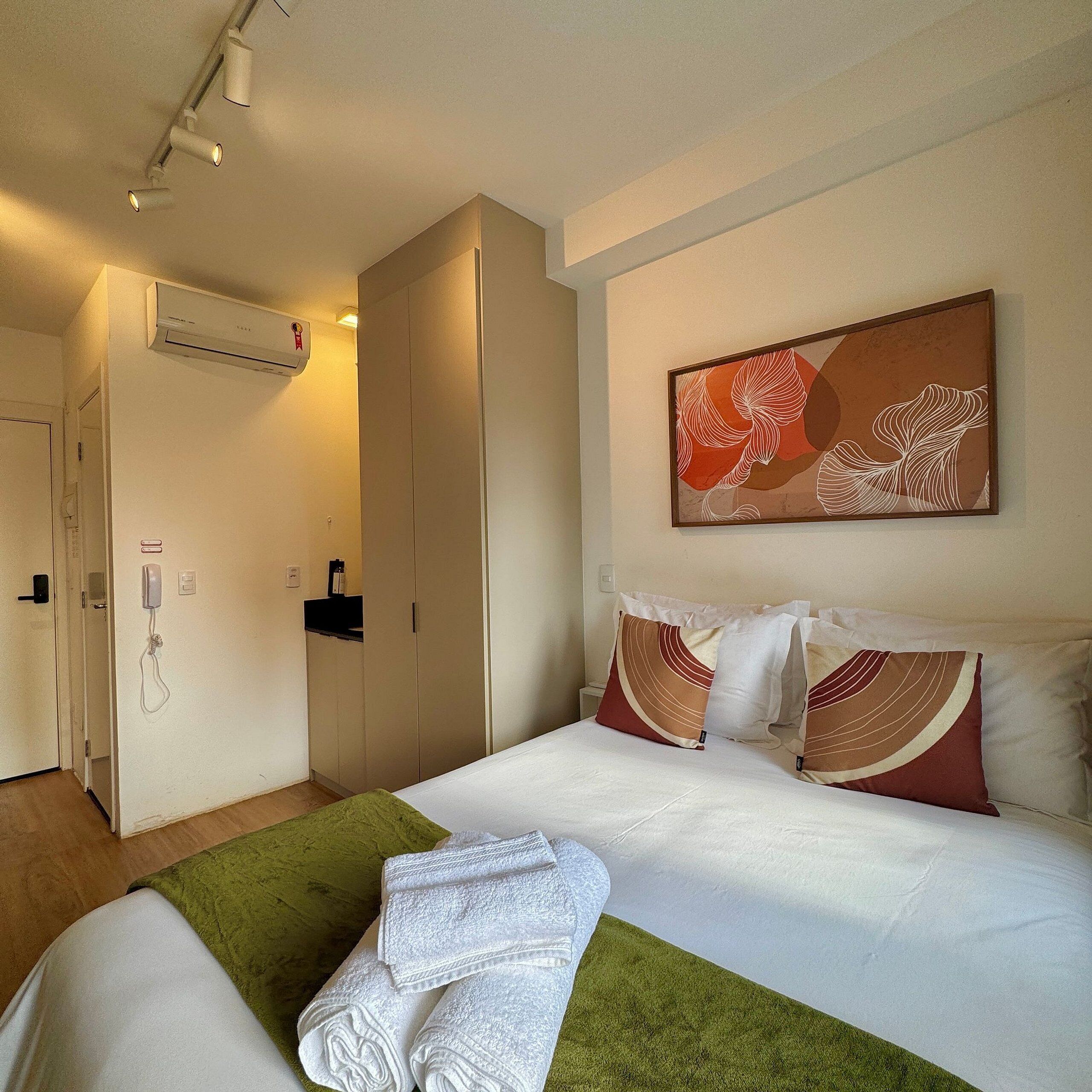Een hotelkamer met een bed, een groene plaid, witte handdoeken, een schilderij aan de muur en een kledingkast.