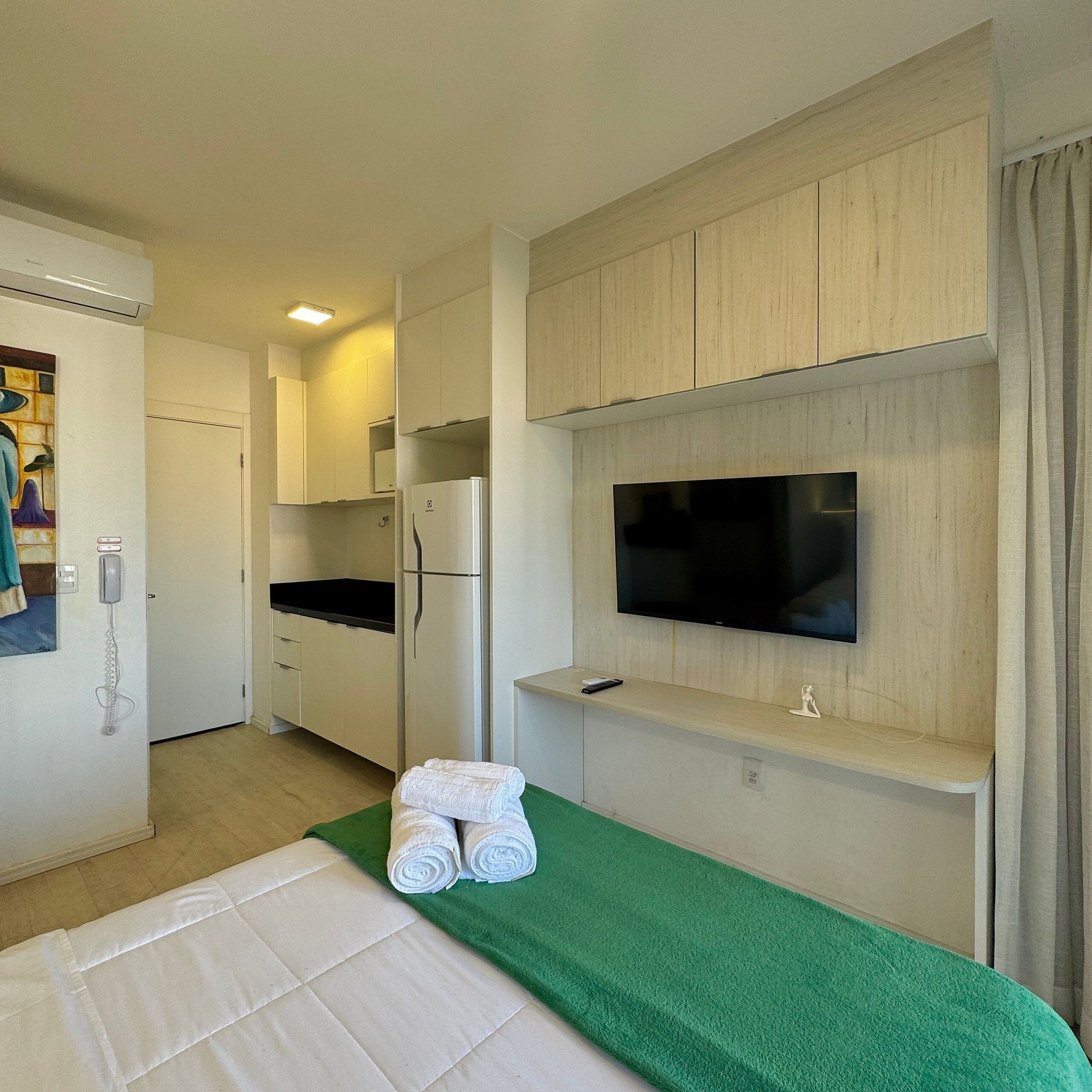 Une chambre d’hôtel avec un lit, un téléviseur fixé au mur, un réfrigérateur, un téléphone et une serviette verte sur le lit.