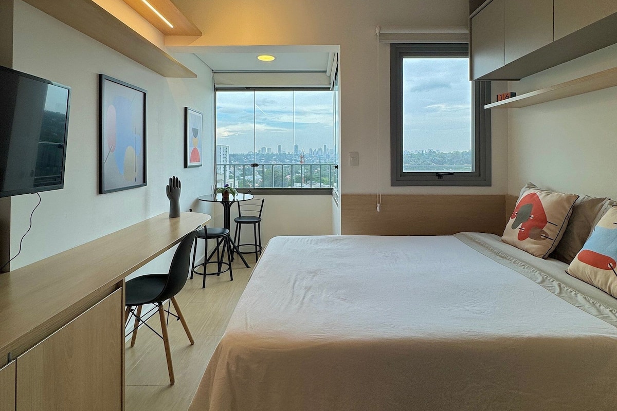 A modern hotel room with a large bed, a desk with chairs, a television, and a city view through the window.