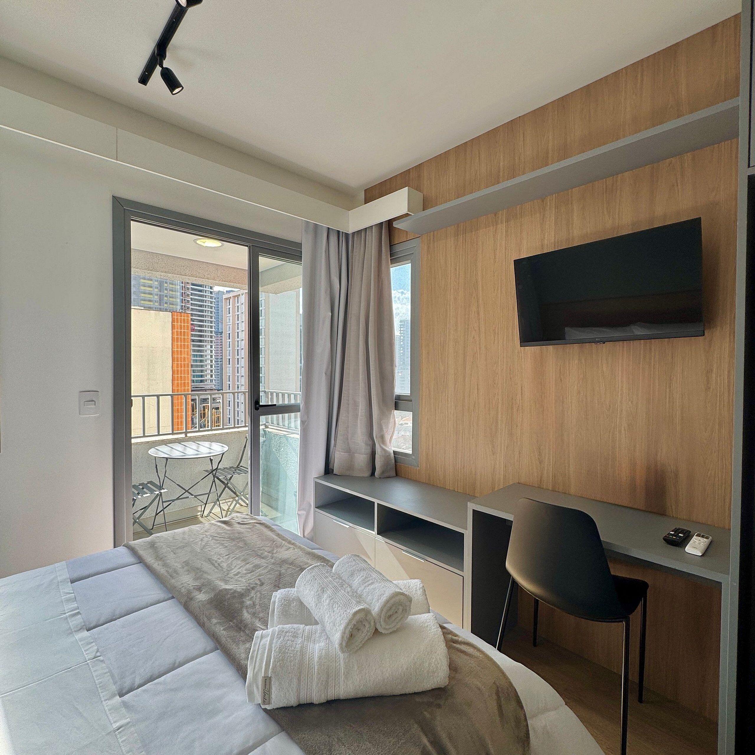 A modern hotel room with a bed, a desk, a chair, a TV, and a balcony with a view of buildings.