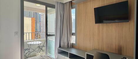 A modern hotel room with a bed, a desk, a chair, a TV, and a balcony with a view of buildings.