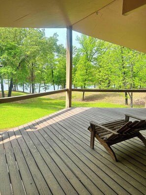 Eine Holzterrasse mit Tisch und Stühlen, die auf eine Wiese mit Bäumen und ein Gewässer blickt.