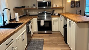 Una cucina moderna con mobili bianchi, un piano di lavoro in legno, un forno in acciaio inox e un forno a microonde. Sono presenti lampade a sospensione e una finestra che permette l'illuminazione naturale.