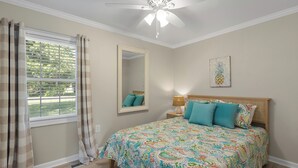 Un dormitorio con cama, ventilador de techo, ventana con cortinas, espejo y una obra de arte con motivos de piña en la pared.