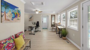 Un interior de hogar con un pasillo que conduce a una puerta, un ventilador de techo, una caminadora, un comedor con mesa y sillas, y un ventanal con persianas.
