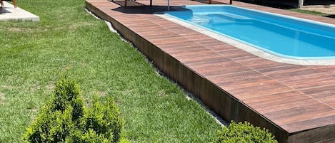A swimming pool with a wooden deck, two lounge chairs, a blue umbrella, and a stone wall in the background.