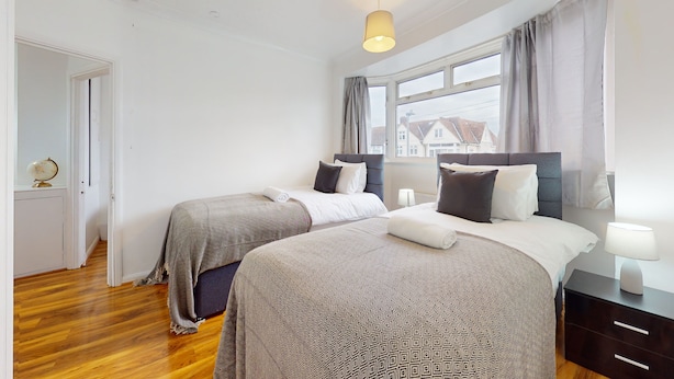 A hotel room with two beds, a wooden floor, a window with curtains, a bedside table with a lamp, and a globe on the wall. - Dagenham - 4 Beds - Sleeps 8 - Wifi (Dagenham)
