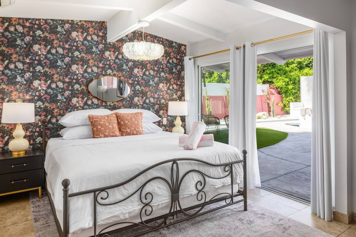 A bedroom with a floral wallpaper, a bed with white bedding, a mirror, and a view of a garden through an open door.