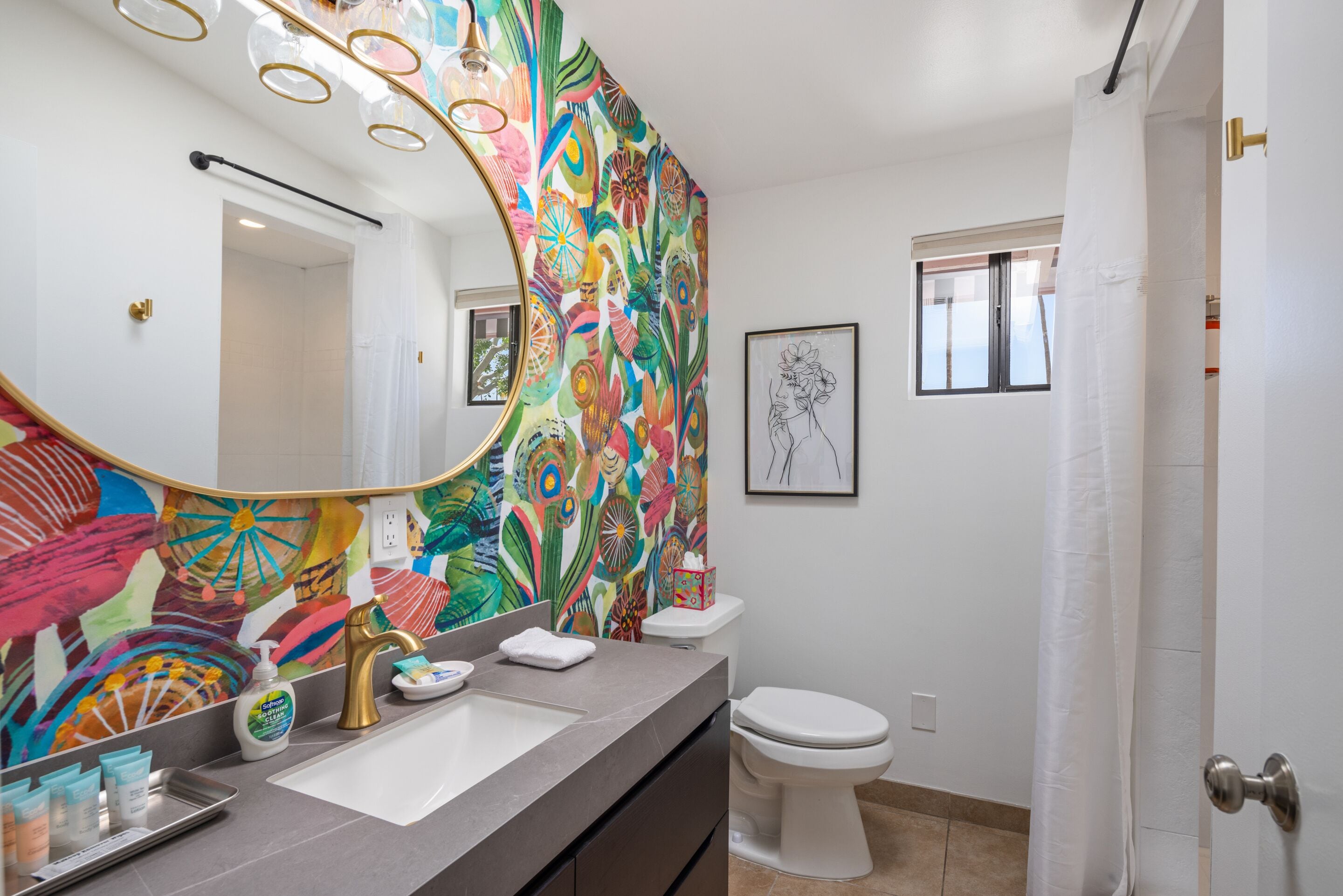 Un baño con un mural abstracto y colorido, un lavamanos moderno con grifo dorado y un inodoro blanco.
