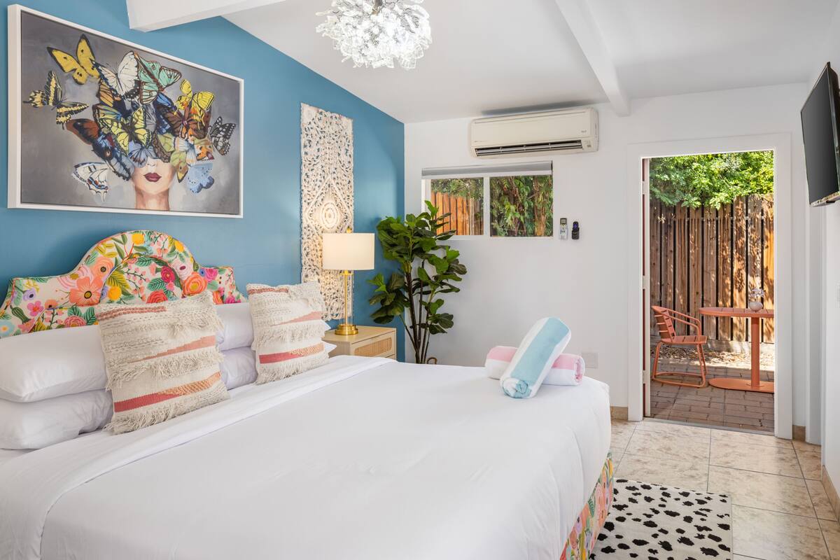 A bedroom with a bed, pillows, a nightstand, a lamp, a plant, and a view of the outside.