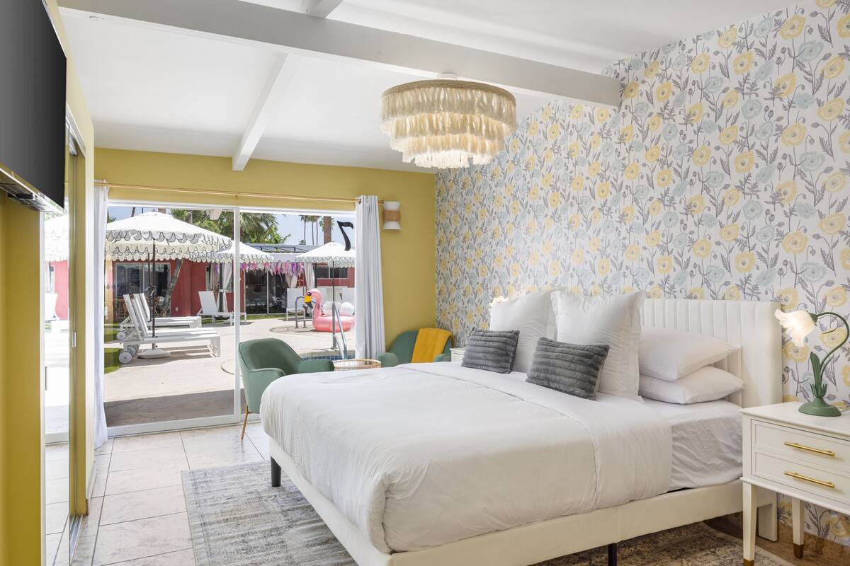 A bedroom with a large bed, a chandelier, and a view of an outdoor area with umbrellas and chairs.