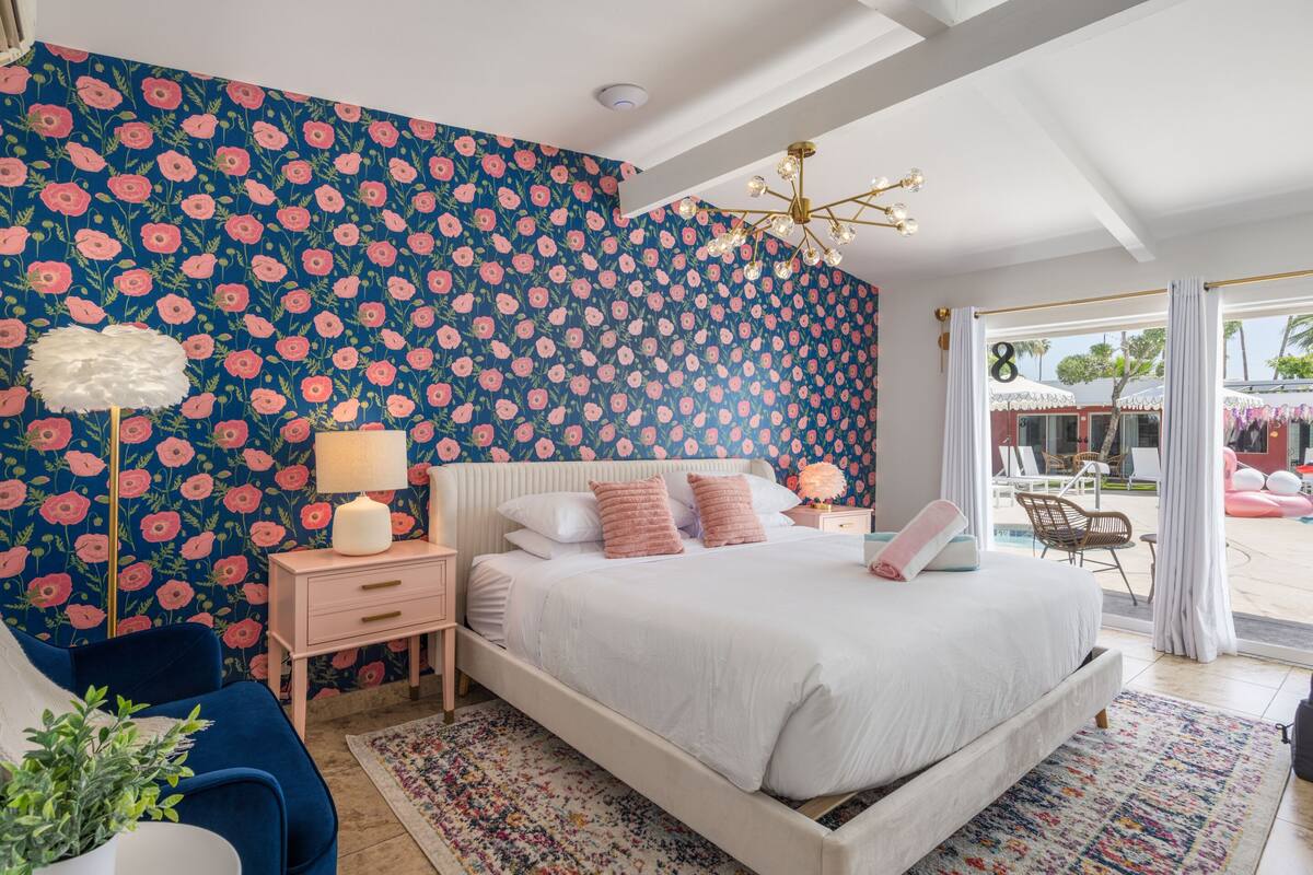 A bedroom with a large bed, a nightstand, a lamp, a chair, and a view of the outside.