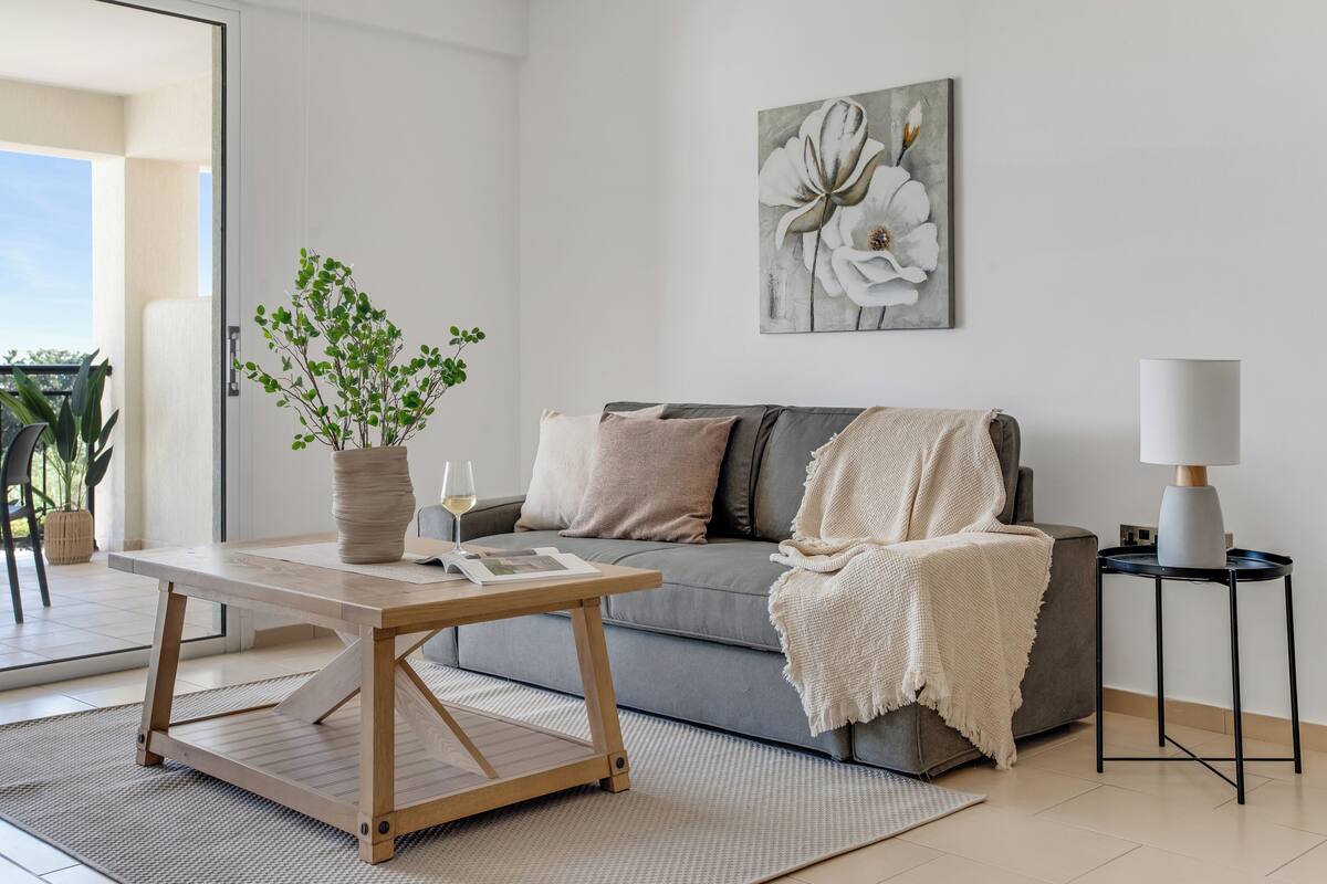Ett modernt vardagsrum med en grå soffa, ett träbord, en krukväxt, en vit lampa och ett stort blommönstrat konstverk på väggen.