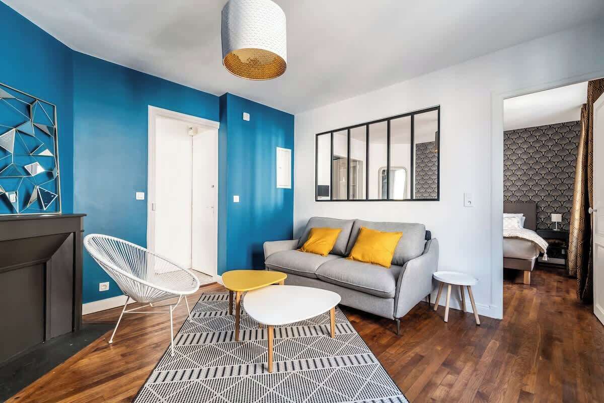 Una sala moderna con una pared azul decorativa, un sofá gris con cojines amarillos, una silla blanca, una mesita pequeña y una alfombra estampada.