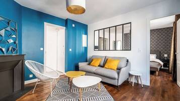 Una sala moderna con una pared azul decorativa, un sofá gris con cojines amarillos, una silla blanca, una mesita pequeña y una alfombra estampada.