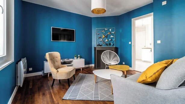 A living room with blue walls, a fireplace, a round coffee table, and a sofa with a yellow pillow. - MM Suites Cathedrale (Reims)