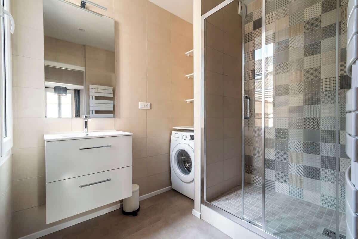 A modern bathroom with a built-in washing machine, a glass shower enclosure, and a large mirror.