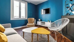 A room with a blue wall, a white sofa, a yellow table, a white chair, a TV, and a white cabinet. - MM Suites Cathedrale (Reims)