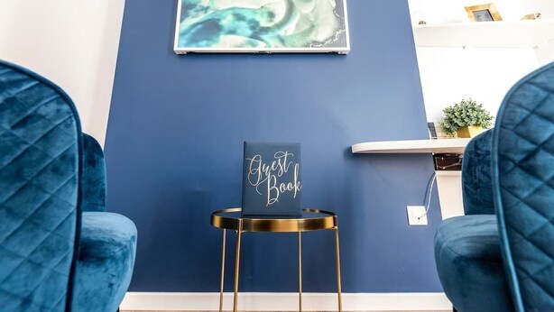 A room with a blue wall, a framed artwork, a 'Guest Book' sign, and two blue upholstered chairs. - MM Suites Marlot (Reims)