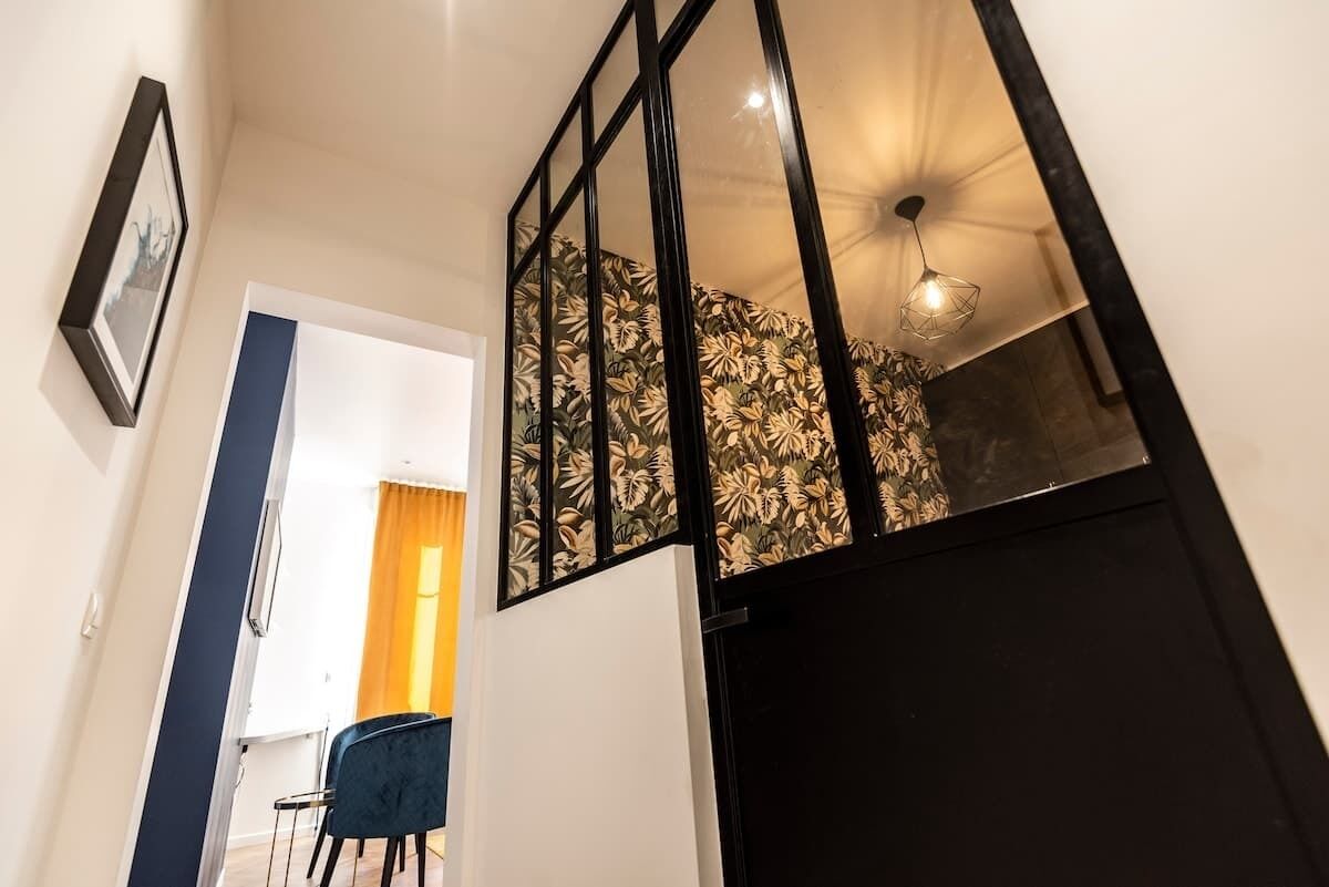 Un couloir avec un miroir à cadre noir, un tableau encadré accroché au mur et une chaise bleue.