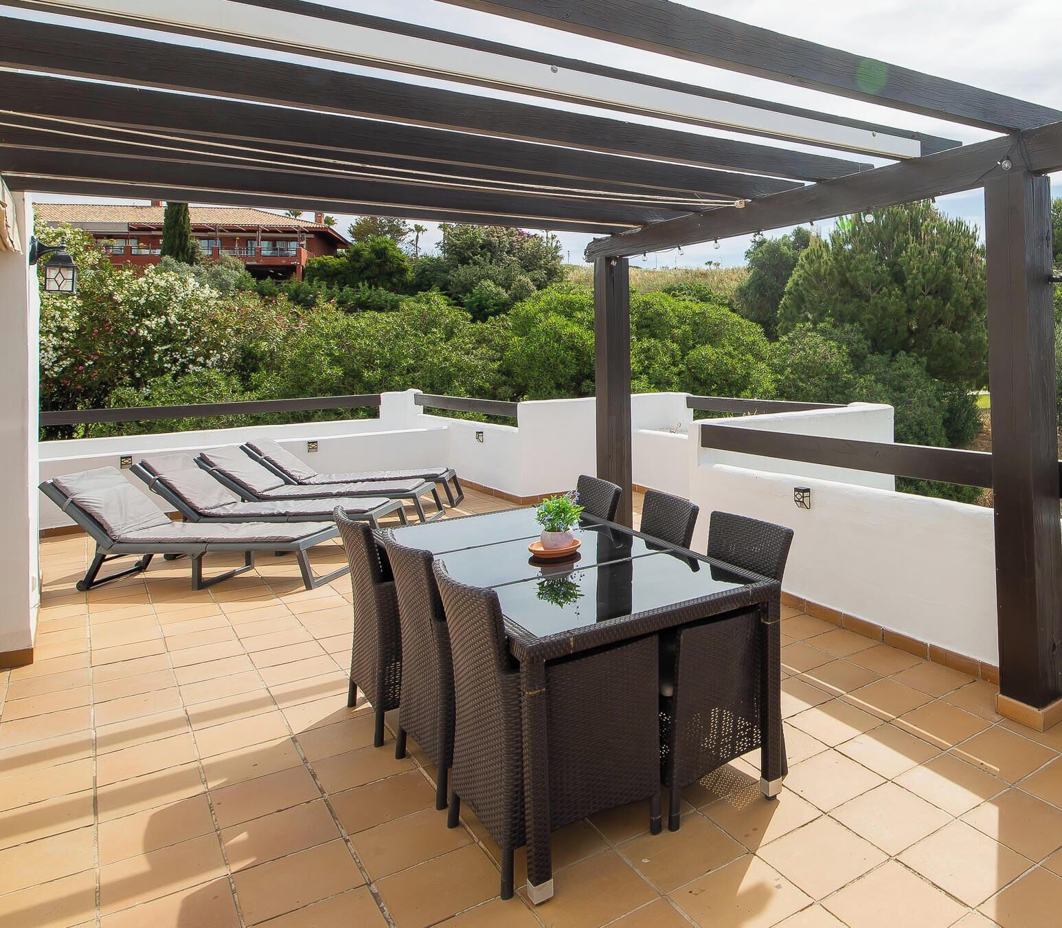 A rooftop terrace with a table and chairs, a pergola, and a view of greenery.